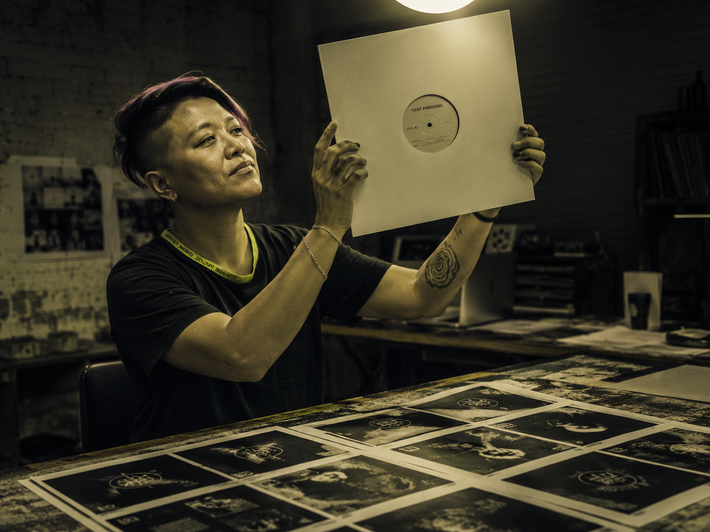 Hands holding 12-inch vinyl test pressing up to studio light, contact sheet of album cover variants on table foreground